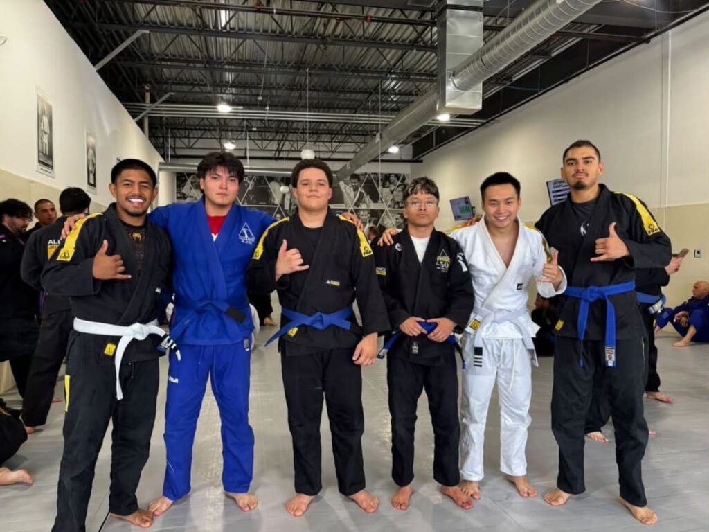 Six young men in mixed gis stand arm-in-arm giving shaka signs after belt promotions