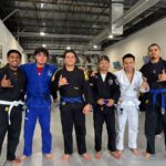 Six young men in mixed gis stand arm-in-arm giving shaka signs after belt promotions
