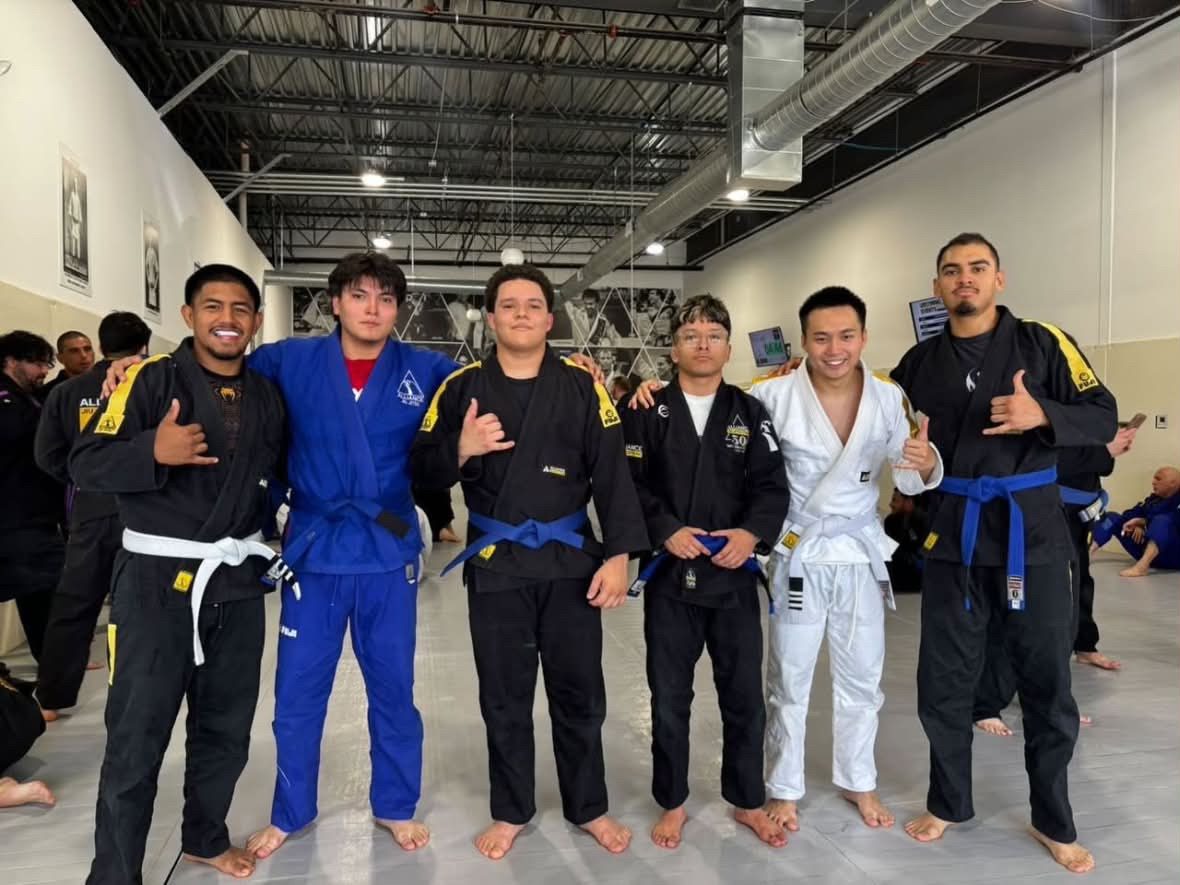 Six young men in mixed gis stand arm-in-arm giving shaka signs after belt promotions