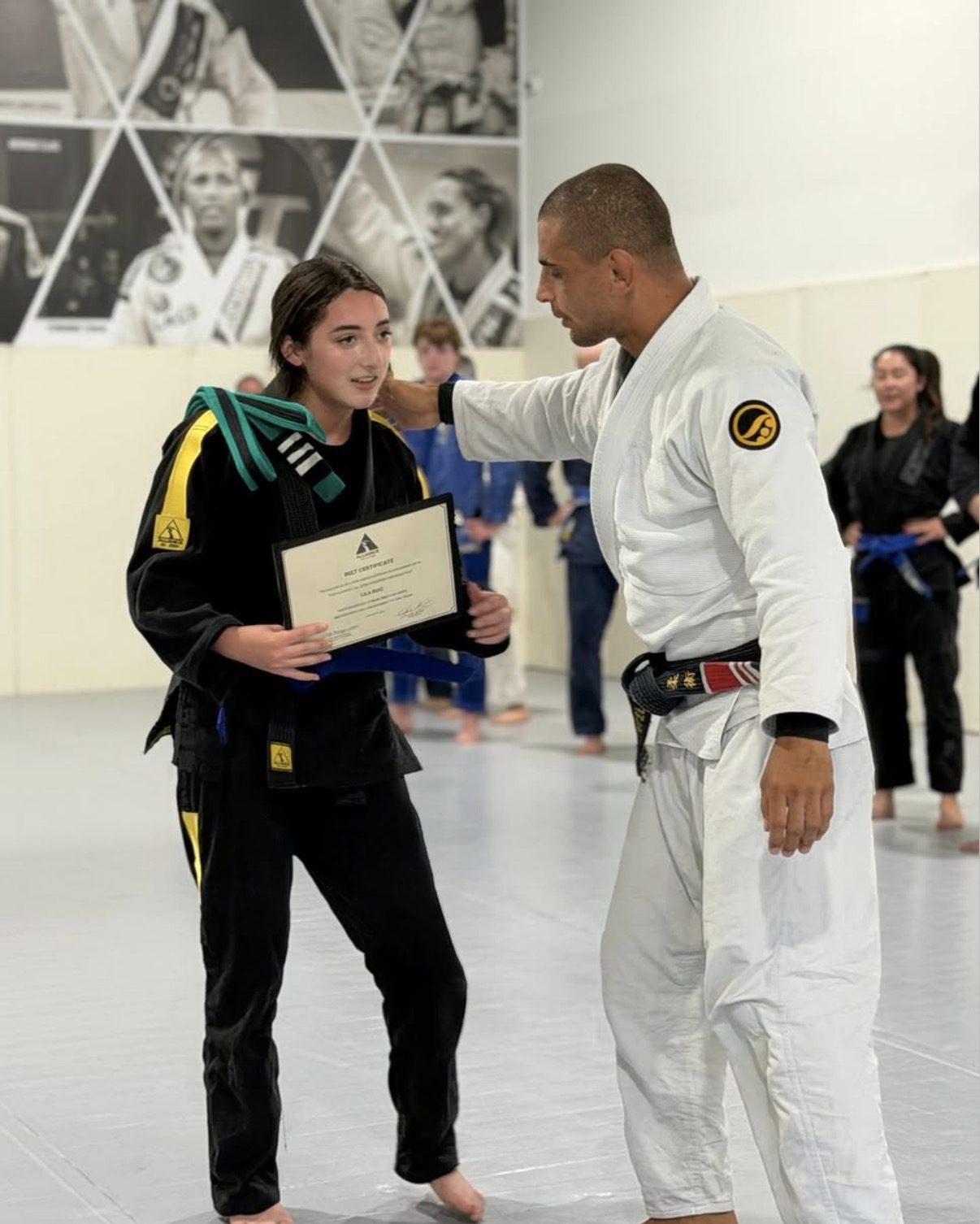 Teen in a black gi holds her blue belt certificate with her coach
