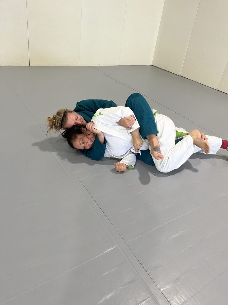 Two women in gis grappling on the mat during a women's jiu-jitsu class