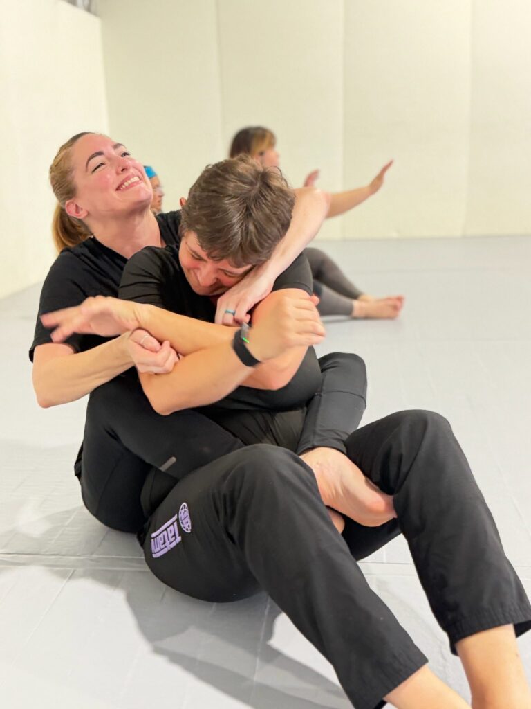 Two smiling students work a seated back-control position during partner drilling