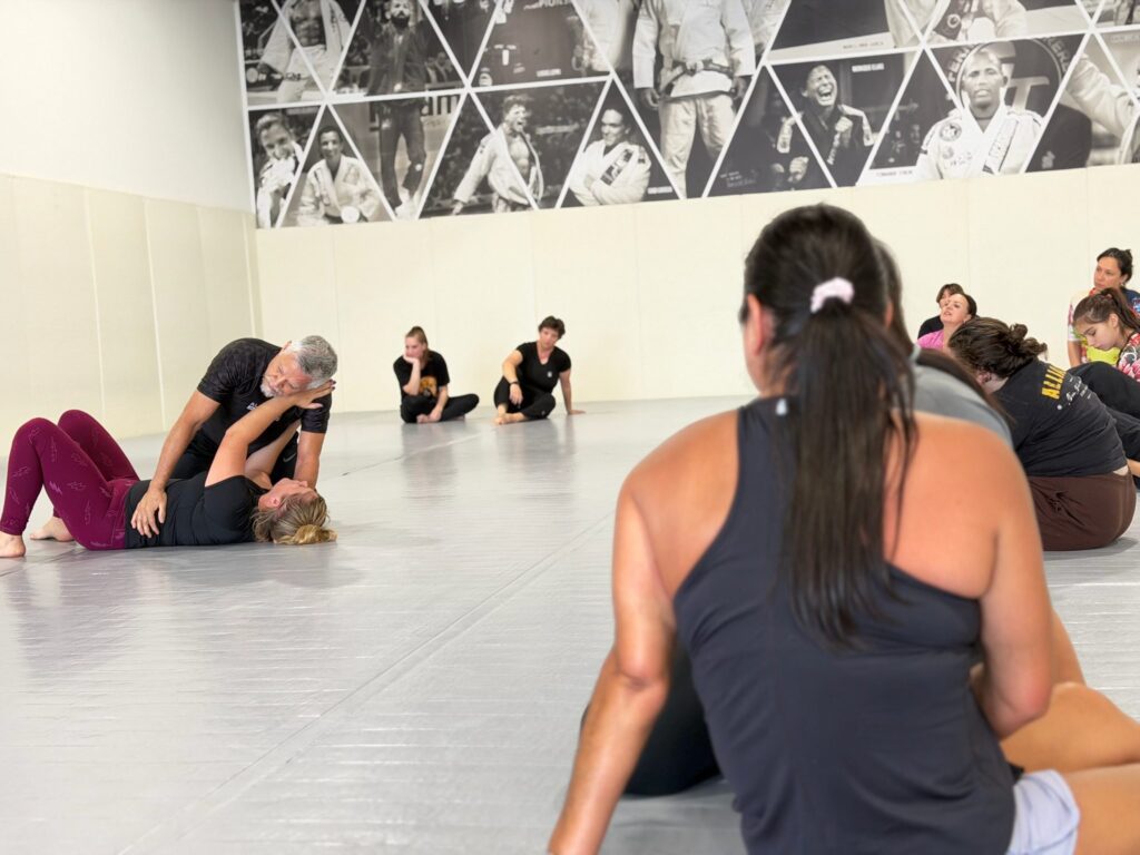 Students watch an Alliance instructor demonstrate a self-defense escape from the mat