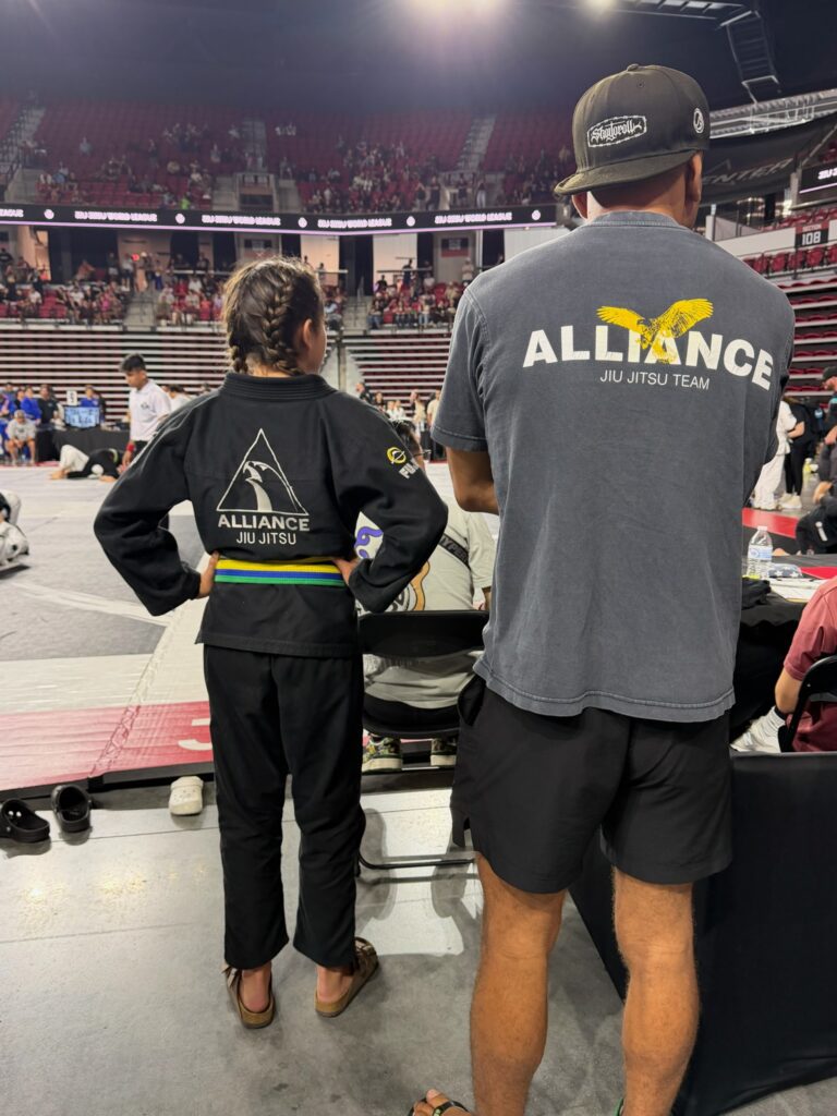 Coach in an Alliance Jiu Jitsu Team shirt waits mat-side with a young competitor