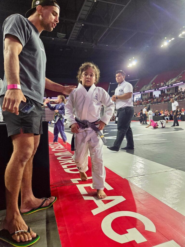 Alliance coach walks a young grey-belt competitor onto the tournament mat