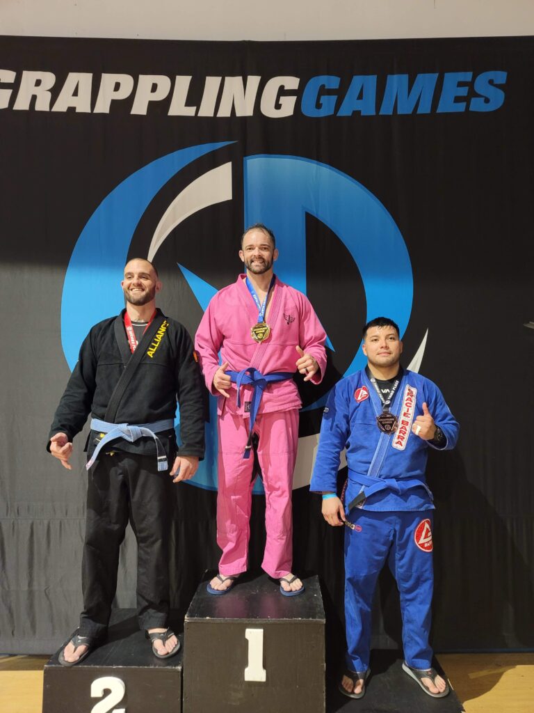 Gold medalist in a pink gi on the top podium step at Grappling Games